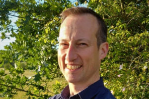White man with short hair smiling on a sunny day in front of green bush