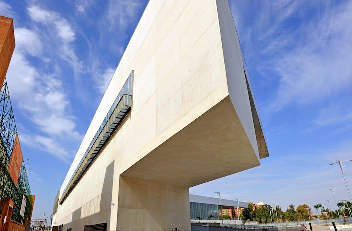 An angle of Fibes exhibition center showing it as a modern architectural building with a unique design. The building features a large, rectangular structure that appears to be cantilevered, extending horizontally without visible support from below. The exterior is made of light-colored concrete or stone, and there is a long horizontal window on the side. The sky is clear with some scattered clouds, and other buildings and structures are visible in the background