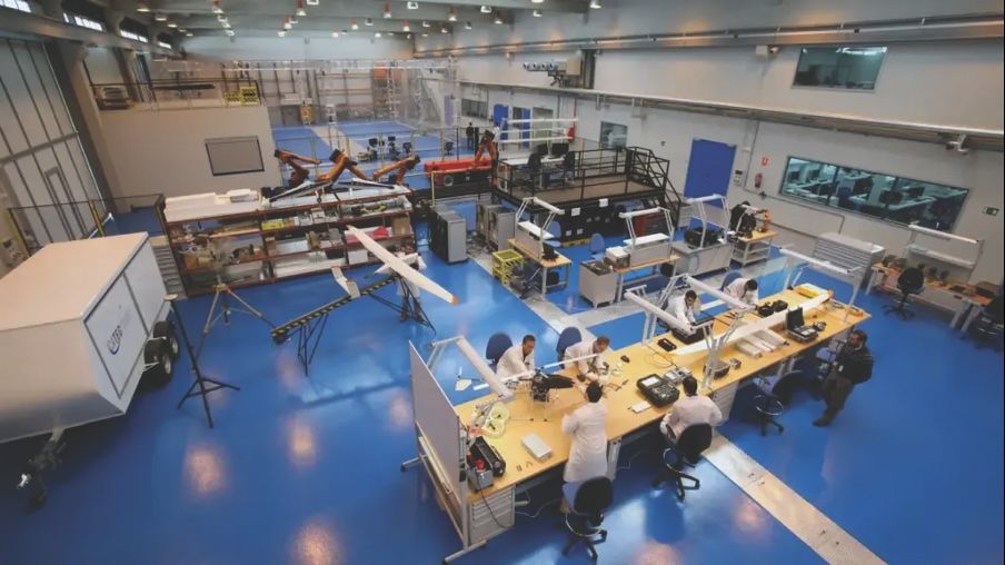 "A large, well-lit laboratory or workshop with several people working at desks. The room has a blue floor and is equipped with various tools, equipment, and workstations. Several unmanned aerial vehicles (UAVs) or drones are visible, including one on a stand and another being worked on by individuals. The space appears organized and designed for research, development, or assembly of UAVs or similar technology."