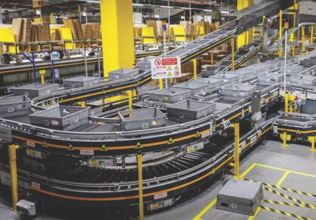 "A large, automated conveyor belt system in a warehouse or distribution centre. The conveyor belts are transporting grey bins, and there are multiple levels of conveyors with yellow structural supports. Various warning signs and safety markings are visible throughout the area, highlighting the importance of safety in this environment."[^i^]
