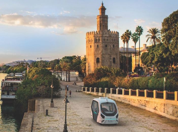 An autonomous vehicle driving along a riverside promenade with the Torre del Oro (Tower of Gold) in Seville, Spain, in the background. The scene includes palm trees, a calm river, and a clear sky during late afternoon or early evening