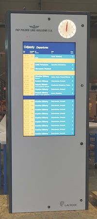  A digital departure board for PKP Polskie Linie Kolejowe S.A., a Polish railway company. The board displays train departure information, including times, destinations, and platform numbers. An analog clock is at the top of the board. 