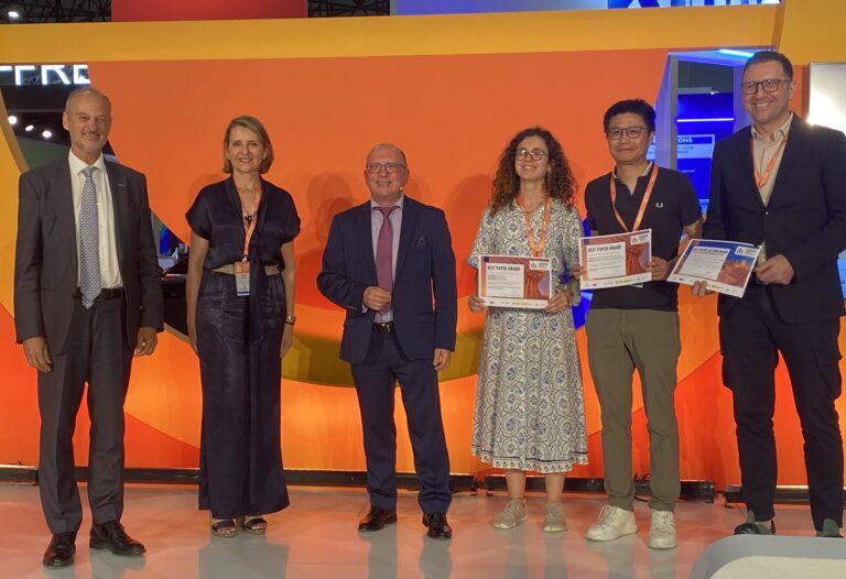 Six individuals standing in a row, some holding certificates, in front of a colorful backdrop with orange, yellow, and blue elements.