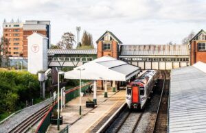 A train station with a train on the tracks, a covered platform, and an overpass connecting two parts of the station. In the background, there are buildings including a tall multi-story structure and one with a large 'T' symbol.