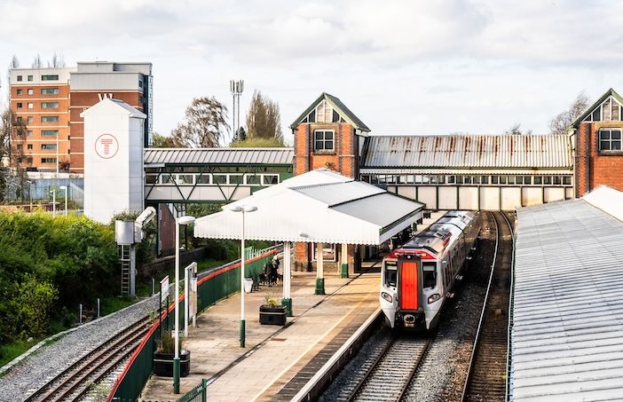 A train station with a train on the tracks, a covered platform, and an overpass connecting two parts of the station. In the background, there are buildings including a tall multi-story structure and one with a large 'T' symbol.