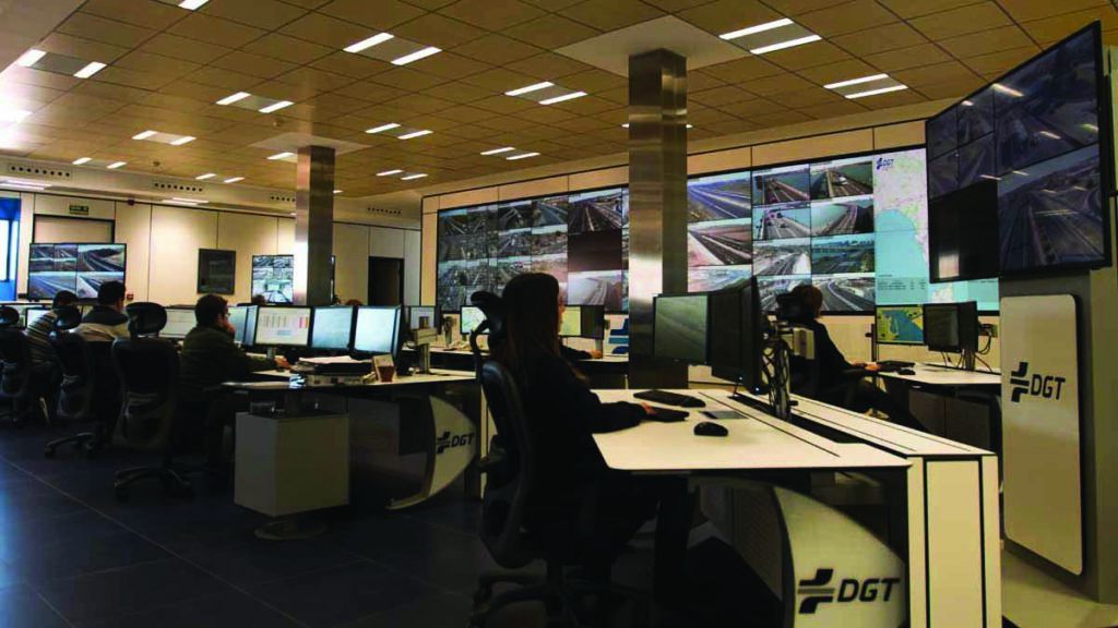 A control room with multiple workstations and large screens displaying various camera feeds. The room is staffed by several individuals working at computers. The workstations and screens are branded with the logo 'DGT,' which stands for Dirección General de Tráfico, the Spanish traffic authority