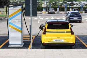 The image shows a yellow Renault electric car parked at a charging station labeled "FREETOX" with the text "ENERGIA 100% GREEN." The car is connected to the charging station via a cable. In the background, there are other cars parked and solar panels installed above some of the parking spaces. This image highlights the use of renewable energy for electric vehicle charging, showcasing advancements in sustainable transportation infrastructure.
