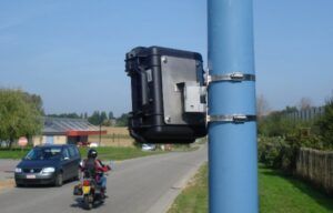 The image shows a black rectangular device mounted on a blue pole using metal straps. In the background, there is a road with a motorcyclist riding away from the camera, and a car parked on the side of the road. The setting appears to be in a rural area with fields and some buildings visible in the distance. This image likely depicts a traffic monitoring or speed enforcement camera setup