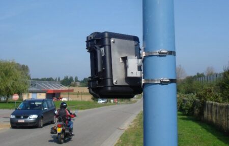 The image shows a black rectangular device mounted on a blue pole using metal straps. In the background, there is a road with a motorcyclist riding away from the camera, and a car parked on the side of the road. The setting appears to be in a rural area with fields and some buildings visible in the distance. This image likely depicts a traffic monitoring or speed enforcement camera setup