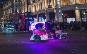 a pedicab (also known as a rickshaw or cycle taxi) on what appears to be a London street at night. The pedicab is illuminated with colorful LED lights - pink, purple, and blue - making it quite eye-catching against the evening backdrop. There are red double-decker buses and classical architecture visible in the background, which is typical of central London areas like the West End