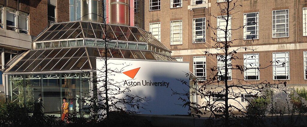 The image shows a building with a sign that reads "Aston University." It features a glass structure with metal pipes on the roof and several windows, and there are trees in the foreground.
