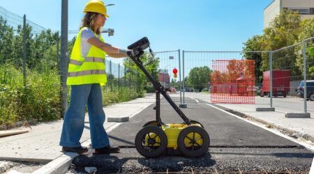 A female worker in yellow hard hat and high-visibility vest operates a yellow wheeled ground-penetrating radar scanner on a paved surface at what appears to be a construction site, with orange safety fencing and barriers visible in the background