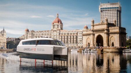 A hydro foiling ferry rises above the water on a river in Mumbai with a backdrop of historical buildings