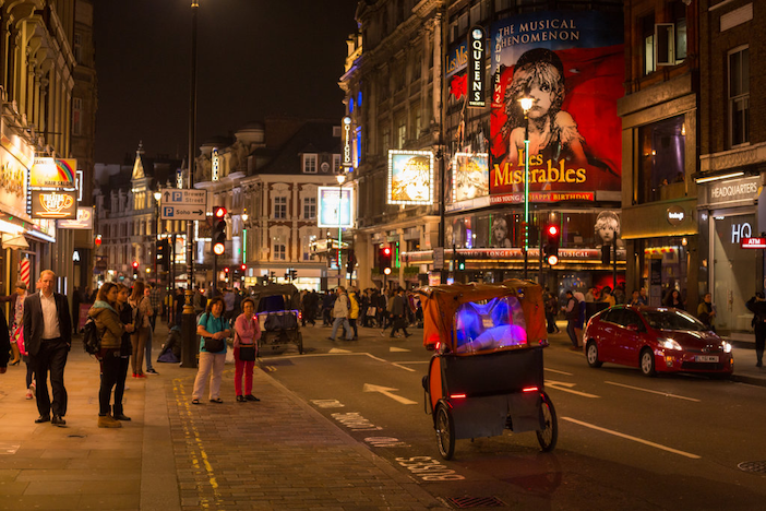 Nighttime city street scene in a theater district with pedestrians, illuminated advertisements including a large 'Les Misérables' poster, a pedicab rickshaw with blue lights is in the foreground, and passing cars