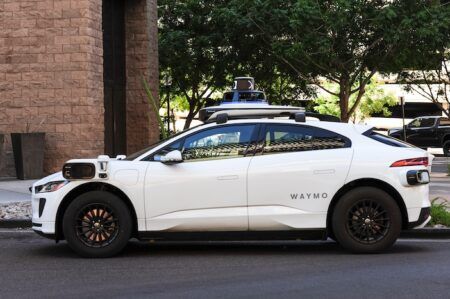 White Waymo autonomous vehicle with sensor equipment mounted on the roof parked on a city street next to a brick building with trees in the background.