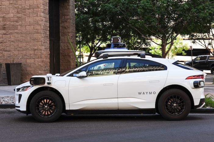 White Waymo autonomous vehicle with sensor equipment mounted on the roof parked on a city street next to a brick building with trees in the background.