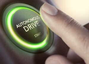 Close-up of a person's finger pressing a circular button labeled 'AUTONOMOUS DRIVE' with 'START' below it. The button is illuminated with a glowing green ring, suggesting activation