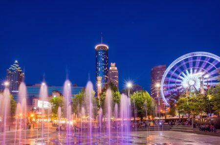 Illuminated water fountains with colorful lighting display at night in downtown city center, with tall buildings and Ferris wheel visible in background against blue twilight sky