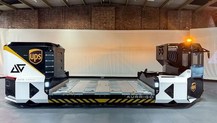 A sleek white and black autonomous cargo dolly vehicle with UPS branding and Aurrigo labeling, featuring a low-profile flatbed design with roller surfaces for baggage handling, displayed in an industrial warehouse setting with exposed brick walls