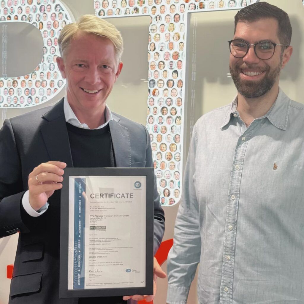 Two professional men smiling and posing together in an office setting, with one holding a framed certificate. The background shows a wall display of employee headshot photos. Both men are dressed in business casual attire - one in a dark blazer and the other in a light blue button-down shirt.