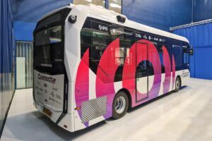 A modern autonomous bus with a distinctive white exterior and large "imo" branding in pink and purple gradient colors, featuring large windows and displaying "Connector" and "Cambridge Biomedical Campus" destination information, housed in a blue-walled testing facility.