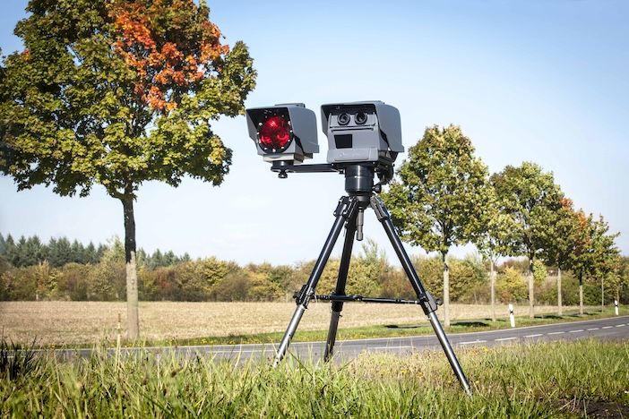 A speed camera enforcement device mounted on a black tripod positioned beside a rural road. The camera unit consists of two white rectangular boxes - one containing a red circular flash unit and another with dual camera lenses. The device is set up on grass alongside an asphalt road, with autumn trees showing orange and green foliage in the background under a clear blue sky. The scene appears to be in a countryside or suburban setting with fields and mature trees visible