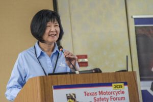 A smiling Asian woman with short black hair stands at a wooden podium, speaking into a microphone. She's wearing a light blue button-down shirt and appears to be giving a presentation. The podium has a banner that reads "2025 Texas Motorcycle Safety Forum" in red text. The setting appears to be an indoor conference or meeting room with beige walls