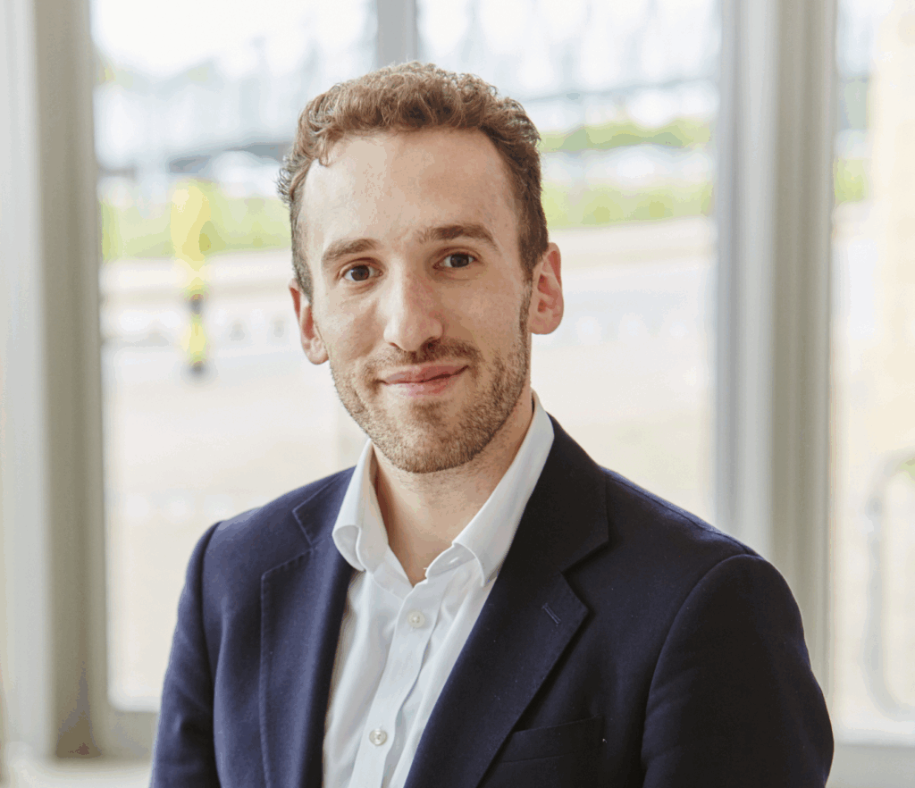 A professional headshot of a man with curly brown hair and light stubble, wearing a dark navy blazer over a white open-collared shirt. He is pictured from the chest up, smiling slightly, with large windows and a bright, softly lit interior visible in the background.