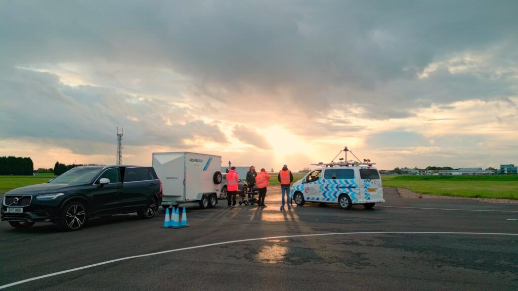 Group of people gathered around vehicles including a vintage blue van on an airfield or large open area during sunset, with dramatic cloudy sky and golden light in the background.RetryClaude can make mistakes. Please double-check responses.