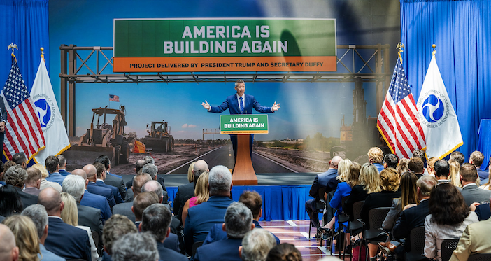 A speaker in a dark suit stands at a podium with 'AMERICA IS BUILDING AGAIN' signage, addressing a seated audience in what appears to be an official government event space. Behind the speaker is a large backdrop displaying construction equipment and infrastructure, with a banner reading 'PROJECT DELIVERED BY PRESIDENT TRUMP AND SECRETARY DUFFY.' American flags and what appear to be departmental flags flank the stage. The audience consists of formally dressed attendees seated in blue chairs, with blue curtains visible along the walls of the venue.