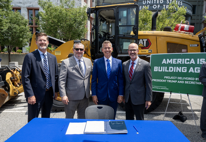 Four men in business suits stand behind a blue table with documents at what appears to be a groundbreaking or construction ceremony. Behind them is yellow Caterpillar construction equipment and a green sign reading 'AMERICA IS BUILDING AG' with 'PROJECT DELIVERED BY PRESIDENT TRUMP AND SECRETAR...' visible. The setting appears to be outdoors in an urban area with buildings and trees in the background