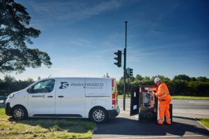 A Swarco worker in an orange high-visibility suit and white helmet is servicing a traffic control box on the side of a road