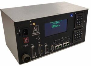 Electronic device with black metal housing. Features a digital LCD display screen showing blue text, multiple input/output ports and connectors on the front panel, numerical keypad on the right side, and various control buttons and indicators. The device is rack-mountable