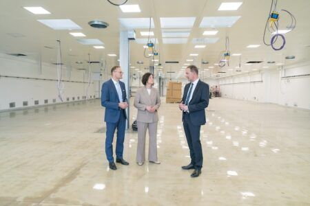 Business executives tour a modern cleanroom or laboratory facility. Three professionals in business attire - one in a blue suit, one in a gray pantsuit, and one in a dark suit - stand in conversation within a large, pristine white space featuring specialized ceiling-mounted equipment with blue and yellow components, likely utility connections or service panels. The facility has polished floors, bright LED lighting, and glass partition walls, suggesting a high-tech manufacturing, pharmaceutical, or research environment. The executives appear to be conducting a facility walkthrough or inspection.