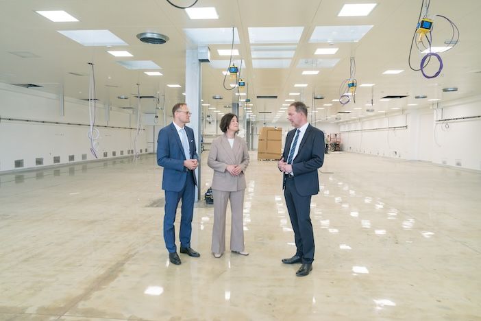 Business executives tour a modern cleanroom or laboratory facility. Three professionals in business attire - one in a blue suit, one in a gray pantsuit, and one in a dark suit - stand in conversation within a large, pristine white space featuring specialized ceiling-mounted equipment with blue and yellow components, likely utility connections or service panels. The facility has polished floors, bright LED lighting, and glass partition walls, suggesting a high-tech manufacturing, pharmaceutical, or research environment. The executives appear to be conducting a facility walkthrough or inspection.