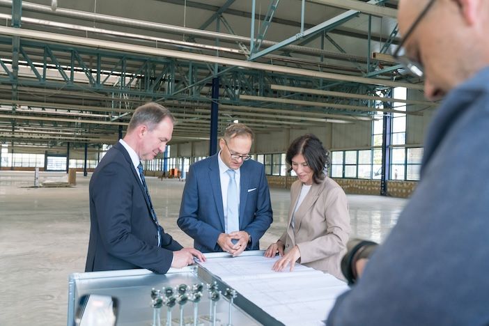 Business professionals examine architectural plans in an industrial warehouse setting. Three people in business attire - one in a black suit, one in a blue suit with glasses, and one in a beige blazer - lean over a table reviewing documents and blueprints. The setting shows a large empty warehouse space with exposed metal ceiling trusses and industrial lighting. A fourth person is partially visible in the foreground.