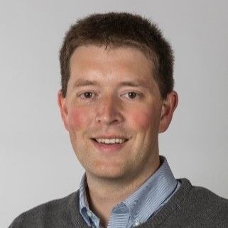 Professional headshot of a young man with short dark hair, smiling at the camera against a plain gray background. He is wearing a gray sweater over a light blue collared shirt