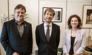 Three professional people standing together in an office setting with framed photographs visible on the wall behind them. From left to right: a man in a dark blazer over a brown shirt; a man in a dark suit with a striped tie; and a woman in a light-colored blazer over a dark top. All three are smiling at the camera.