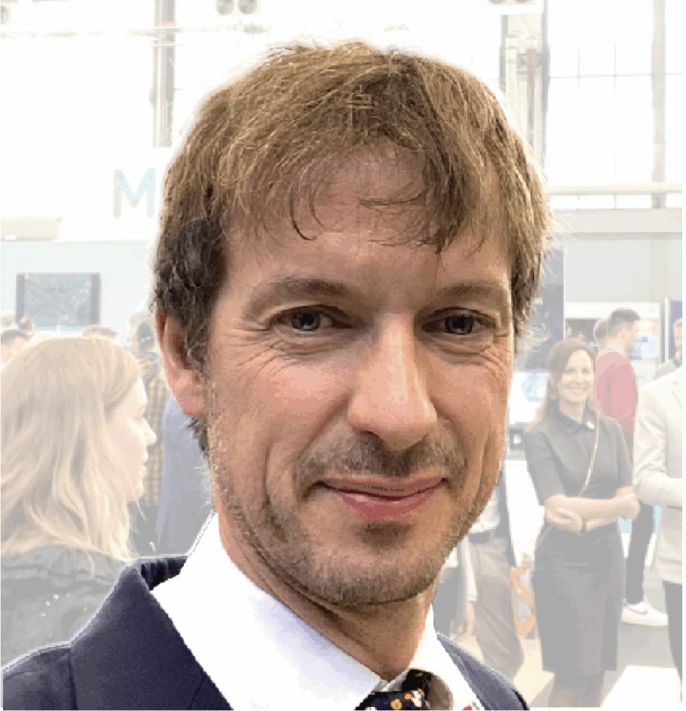 "Professional headshot of a man with short blonde hair and facial stubble, wearing a dark suit jacket and white shirt with a patterned tie, smiling at the camera. The background shows a blurred indoor venue with other people visible in the distance."