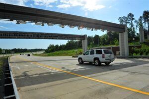 White SUV driving on a multi-lane highway beneath electronic tolling gantries. The road features yellow lane markings and is surrounded by green trees and vegetation under a partly cloudy blue sky. Multiple overhead toll collection structures span the highway, with electronic sensors and equipment mounted on the gantries for automated toll collection.