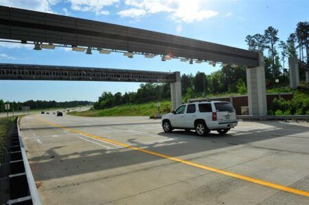 White SUV driving on a multi-lane highway beneath electronic tolling gantries. The road features yellow lane markings and is surrounded by green trees and vegetation under a partly cloudy blue sky. Multiple overhead toll collection structures span the highway, with electronic sensors and equipment mounted on the gantries for automated toll collection.