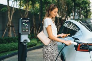 Young woman travel with EV electric car charging in green sustainable city outdoor garden in summer. Urban sustainability lifestyle by green clean rechargeable energy of electric BEV vehicle innards