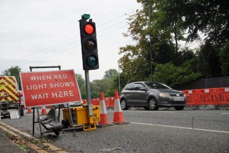A temporary traffic signal showing red at a road construction site, with a warning sign reading "WHEN RED LIGHT SHOWS WAIT HERE" positioned beside traffic cones and barriers as a vehicle passes through the work zone