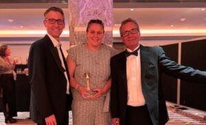 Woman in evening dress holding an award flanked by two men in black tie dinner suits