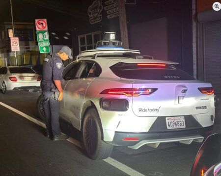A San Bruno police officer peers into a white Waymo autonomous vehicle stopped at night with emergency lights activated. The driverless car, identifiable by sensor equipment on its roof and 'Ride today' branding on its side, was pulled over after making an illegal U-turn during a DUI enforcement operation. The officer stands beside the empty driver's seat examining the vehicle's interior