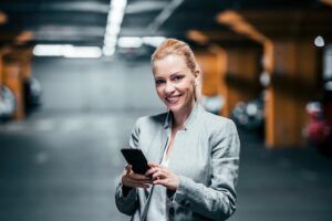 Woman in a grey blazer stands in an underground parking garage, smiling while holding a smartphone in both hands. Orange lighting illuminates the concrete structure behind her.