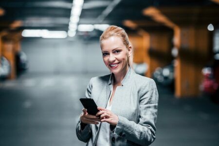 Woman in a grey blazer stands in an underground parking garage, smiling while holding a smartphone in both hands. Orange lighting illuminates the concrete structure behind her.