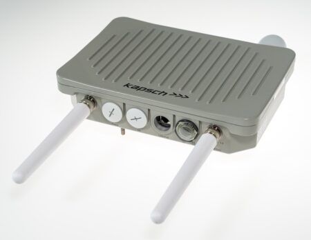 Kapsch RSU is first C-V2X hardware certified under new FCC rules A grey rectangular Kapsch-branded roadside unit with ventilated top casing and two white antennas extending from the front panel. The unit features multiple connection ports including power, I/O, LAN PoE and coaxial connectors along the front edge.