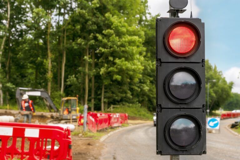 SRL secures long-term contract for remotely operated traffic signals Portable three-aspect traffic signal unit displaying red light at roadworks site, with road barriers, construction equipment and workers visible in background on tree-lined road