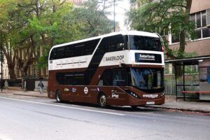 Brown and white liveried Bakerloop bus on London street, showing route map on side panel and modern double-deck design